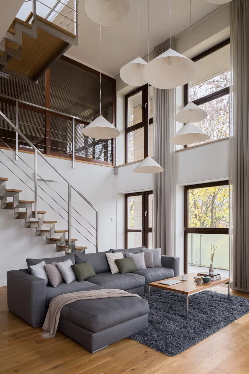LUBA Immobilien UG - Immobilien - Modernes Wohnzimmer mit grauem Ecksofa, Couchtisch aus Holz, großen Fenstern, Hängeleuchten und einer Treppe mit Holzstufen und Metallgeländer.