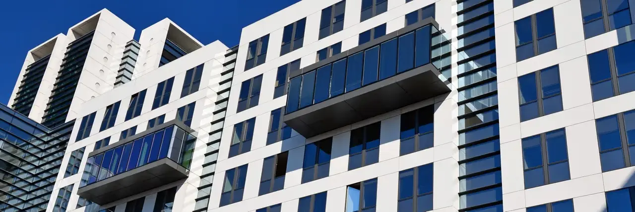 Modern white and blue office building facade with large windows and two glass balconies under a clear blue sky.